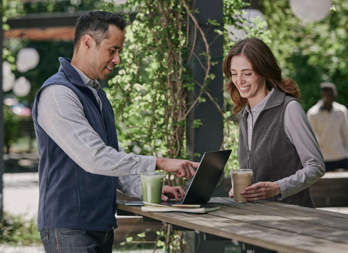 Two people using a laptop outdoors with drinks on a table.
