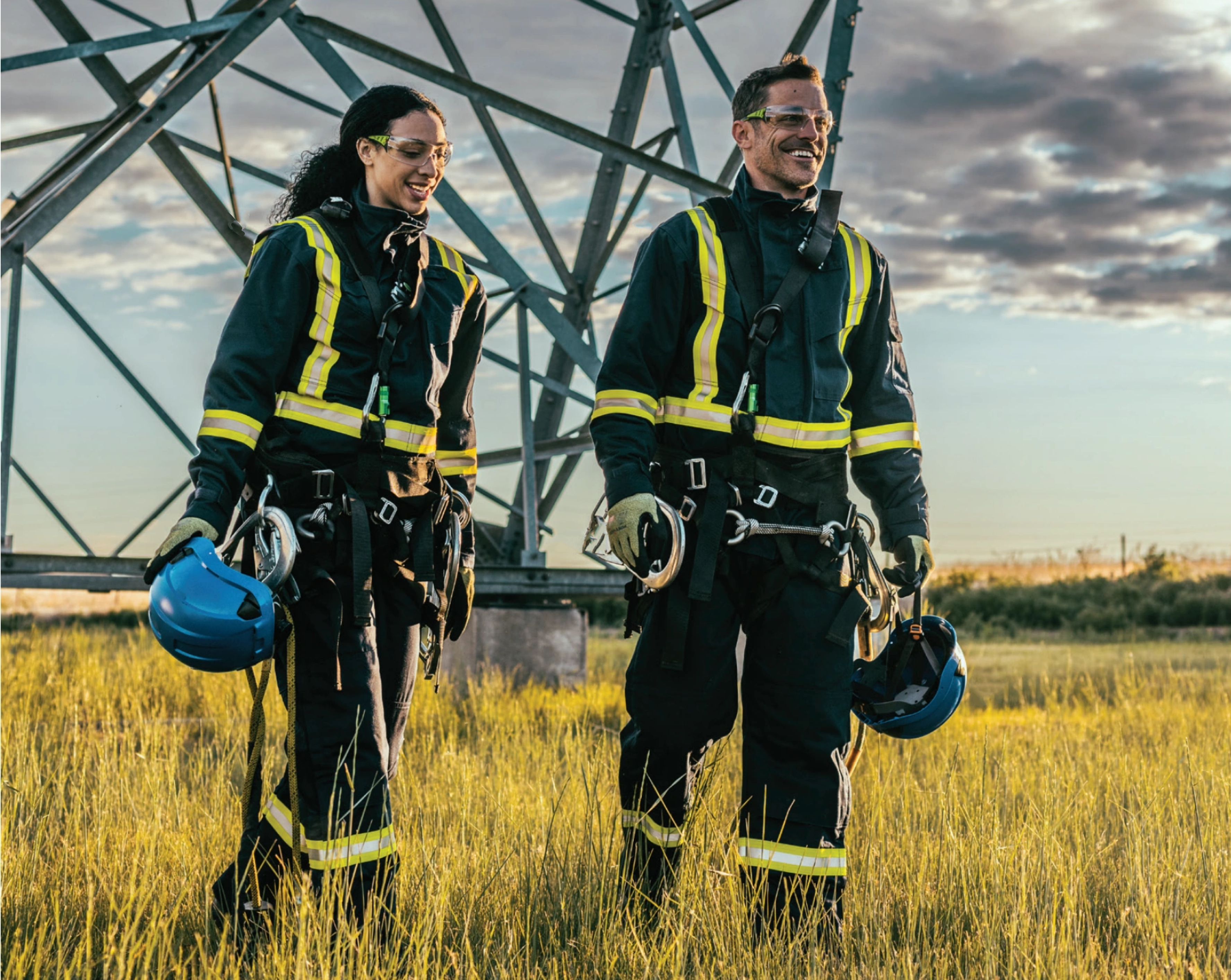 Two individuals in high-visibility safety gear walking through a field with a large metal structure in the background.
