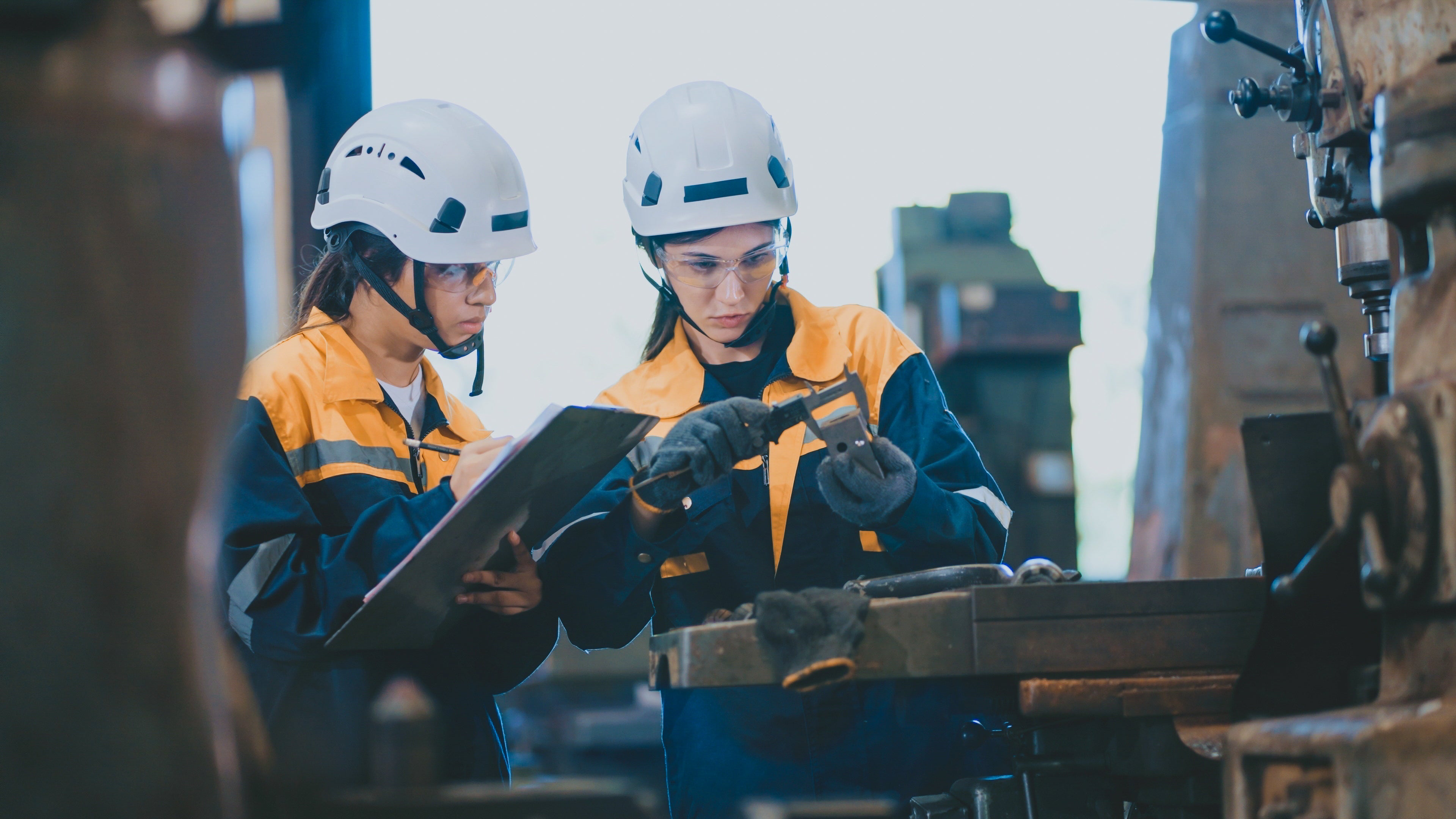 Two workers in a factory setting, one holding a clipboard and the other measuring equipment.
